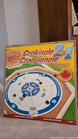 Box front showing the 2-in-1 Crokinole and Checkers game set, with circular Crokinole board and checkers overlay image, Canadian maple leaf symbol indicating wooden game made in Canada, age suggestion 6+
