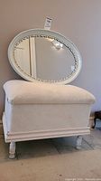 Photo showing the white cushioned stool with a wooden base and the oval mirror resting on it, both with white finishes and floral details on the mirror frame.