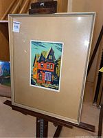 Framed print with beige mat board and metallic frame showing a colorful architectural scene of a red brick house with steep rooflines under blue sky.