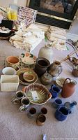 Full view of assortment of stoneware and ceramic pieces on carpet with textiles in background. Includes bowls, pitchers, mugs, small pots.