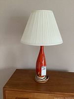 Full view of the lamp showing white pleated shade, glazed ceramic base in bright orange with brown and yellow accent, wired base on wooden platform.