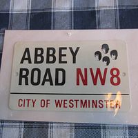 Photo displaying the Beatles Abbey Road NW8 collectible metal sign with City of Westminster text and Beatles silhouettes, on a fabric surface.