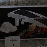 Photo of mandoline slicer on box showing white plastic frame, stainless steel slicing blade, safety handguard, and assorted sliced vegetables on box art.