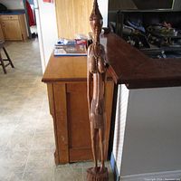 Full side view of the tall wood carving near a kitchen island, showing the full height and side profile.