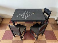 Top and angled view of black children's chalk table with 3 black wooden chairs arranged around it on checkered floor.