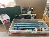 Plastic bin with gift wrapping paper rolls and assorted ribbons, with multiple boxes of NOMA white string lights behind