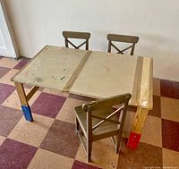 Children's wooden drawing table with three wooden IKEA chairs around it. Table surface shows drawing marks and wear. Color pads on table legs.