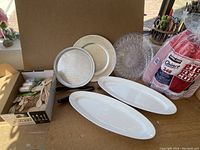Wooden cutlery opened box, red plastic solo cups in original package, white oval ceramic platters, round ceramic and clear decorative plates.