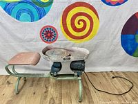 Side view of electric pottery wheel and cushioned seat connected by metal frame, showing power cord and wheel.