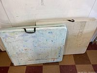 Two folded plastic folding tables leaning against wall. One appears white with some marks, the other light green with paint stains.