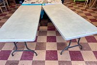 Two large plastic tables side by side, showing rectangular plastic tops with foldable metal legs on tiled floor.