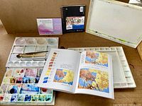 Photo showing multiple watercolour palettes including one with dried paint, paintbrushes, a spiral-bound paper pad, a small box of watercolour paints, and a watercolour paper sheet mounted on a board