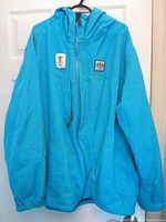 Light blue full-zip men's jacket with hood, Olympic patches on chest, elastic cuffs, and side pockets.