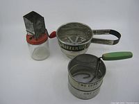Three vintage kitchen gadgets: Androck nut meat chopper, Foley flower sifter with label, and a second flower sifter with green handle.