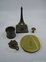 All five vintage metal items displayed together on a white background including a brass compact mirror, metal napkin ring, candle holder, Eiffel Tower figurine, and lion head hook.
