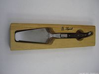 Cake slicer tool resting inside the wooden holder displaying full length of the tool and holder with Laguiole branding.