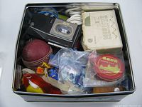 Top view of tin showing a black AVR cassette recorder, vintage electric cloth cutter box, cricket ball, and various small items in plastic bags.