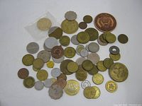 Photo of a large assortment of old coins and tokens laid out on a white surface, showing a variety of metal types including brass, copper, silver, and gold tones, some round others with unique shapes.