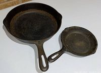 Two cast iron skillets on floor showing top cooking surfaces and handles, visible rust and wear.