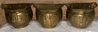 Three brass flower planter pots arranged side by side showing front decorative details and overall shape.