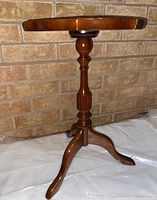 Front view of small wooden side table showing round scalloped top, turned pedestal, and three curved legs against a brick wall.