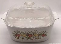 Full view of Corningware Spice of Life 5L casserole dish with clear glass lid in place, showing integrated side handles and vegetable decoration on dish body.