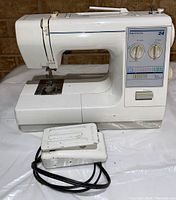 Front view of white Kenmore 24 sewing machine with foot pedal and cut power cord on white surface against brick wall.