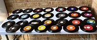 Overview of a lot of forty-five 45 RPM 7 inch vinyl records, displayed on a table. Records have varying center labels and appear in used condition with visible scratches and wear.