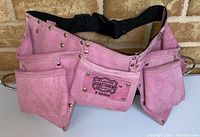 Front view showing pink suede material, multiple tool pockets, adjustable black waist strap, and metal rivets.
