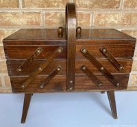 Front view of closed vintage wooden sewing box showing accordion style expanding design and wood handle on top. Angled legs supporting box.