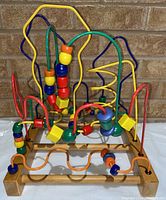 Wooden bead maze toy with multicolored beads on intricately bent metal wire paths and wooden base, angled front view against brick wall.