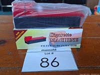 Photo of the cigarette machine on its original box showing the red and black plastic device and box labeled 'Cigarette Machine Filter Tube Injector'