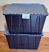 Two stacked black plastic storage bins with gray lids closed, showing ribbed sides and flat lids.