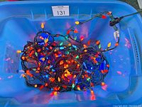 Large blue tote filled with multi-colored outdoor Christmas light strings, plugged in and illuminated, showing red, blue, orange, yellow, and green bulb-shaped lights on green wire.