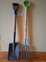 Photo showing the full length of the two short-handled tools leaning against a wall: a flat shovel with rectangular blade and a loading fork with four tines and a green plastic grip.
