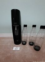 Front view of black SodaStream soda maker with three clear carbonating bottles beside it on carpeted floor.