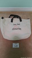 White canvas tote bag with black handles and embroidered phrase displayed on beige carpet, also showing lot number.