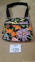 Front view of the Thirty-One floral bag showing colorful 70's themed floral pattern, front zippered pocket, and thick adjustable black strap.