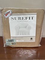 Front view of sealed SureFit chair slipcover packaging, beige color, indicating fit for arm chairs, includes Scotchgard protection.