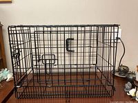 Black collapsible wire metal pet crate placed on wooden surface against wall. Front and side doors visible with latch mechanism and central handle on top.