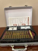 Opened metal briefcase displaying the board game components inside, including instruction sheet, plastic cases with numbers, and play money.