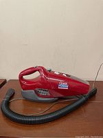 Front and side view of the red Dirt Devil Quick Power handheld vacuum with hose attachment on a wooden surface.