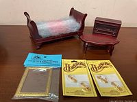 Photo showing the miniature sleigh bed with pastel bedding, small dark wood display cabinet, oval coffee table, and packaged miniature accessories on a wooden surface.