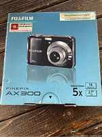 Front of retail box showing Fujifilm FinePix AX300 camera in black, with specifications such as 14 MP, 5x optical zoom, and 2.7-inch LCD screen.