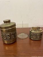 Brass container, clear acrylic holder, brass lighter arranged on wood surface.