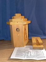 Front view of natural wood bird house with hexagonal body and stepped roof, showing circular entrance hole, perch, and detached base/mounting piece alongside an instruction sheet.