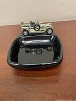 Black ceramic ashtray with a reflective glossy finish, showing the metal vintage car figurine attached on one edge.