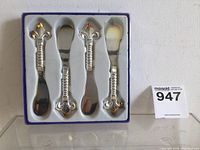Set of four silver-colored butter spreaders with decorative fleur-de-lis handles in a box with foam insert