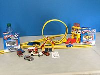 Wide shot of yellow Hot Wheels track setup with loop, race tower, traffic barriers, various loose Hot Wheels cars and two new boxed Metallics cars