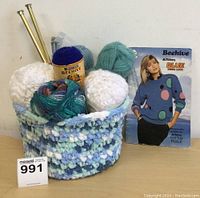 Basket filled with four balls of yarn, two brass needles, and Beehive knitting pattern book.